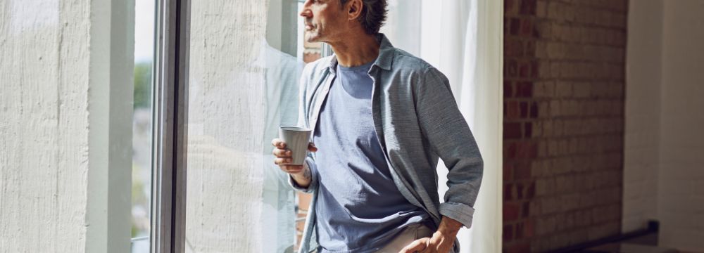 Older man staring out window holding cup of coffee in right hand, leaning on glass