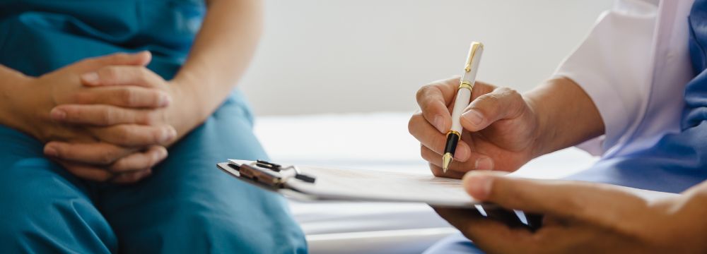 Man sitting on doctor's physical table folding hands in lap white provider takes notes with pen on clipboard